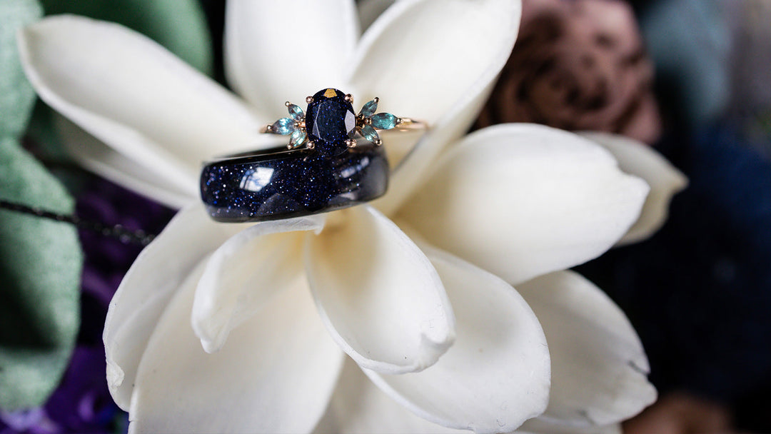 Close-up of a single unique ring featuring a wide blue sandstone band topped with a gold-mounted dark blue oval gem and teal accents, resting on a white flower.