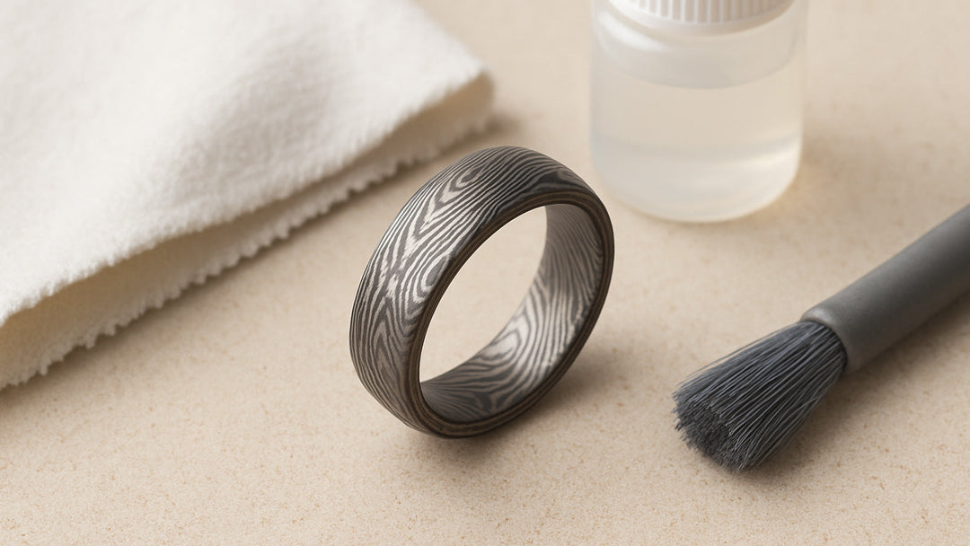 A striking patterned metal ring (Damascus style) is displayed with a white cleaning cloth, liquid, and a brush
