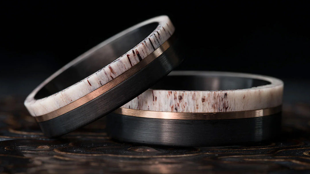 Two black men's rings with antler inlay and rose gold accents on a dark textured surface