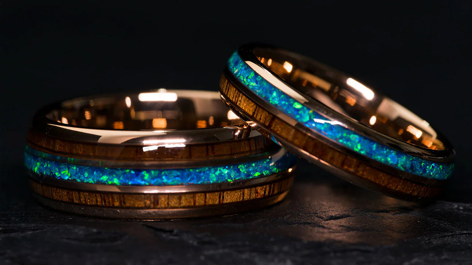Two Sagaband wood and opal inlay bands rest on a dark textured surface