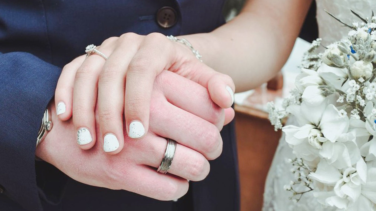 Sagaband textured metal men's wedding band on interlocking couple hands with white bouquet