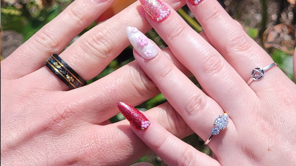 Hands wearing a black wedding ring, engagement ring, and pink floral nail art outdoors.