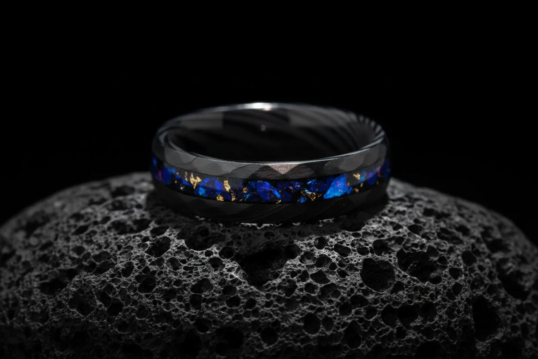 A textured black Sagabands ring with a vibrant blue and gold flake inlay rests atop a porous volcanic rock.