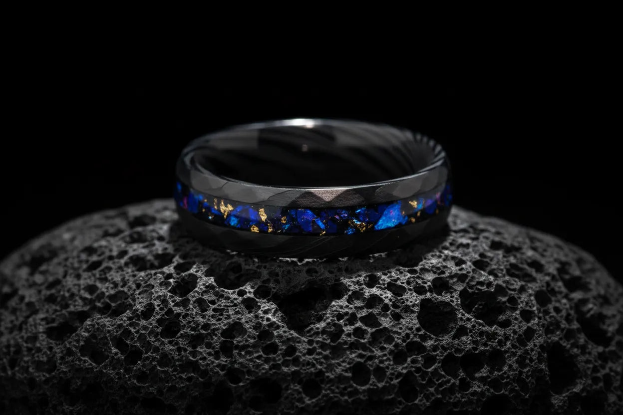 A textured black Sagabands ring with a vibrant blue and gold flake inlay rests atop a porous volcanic rock.