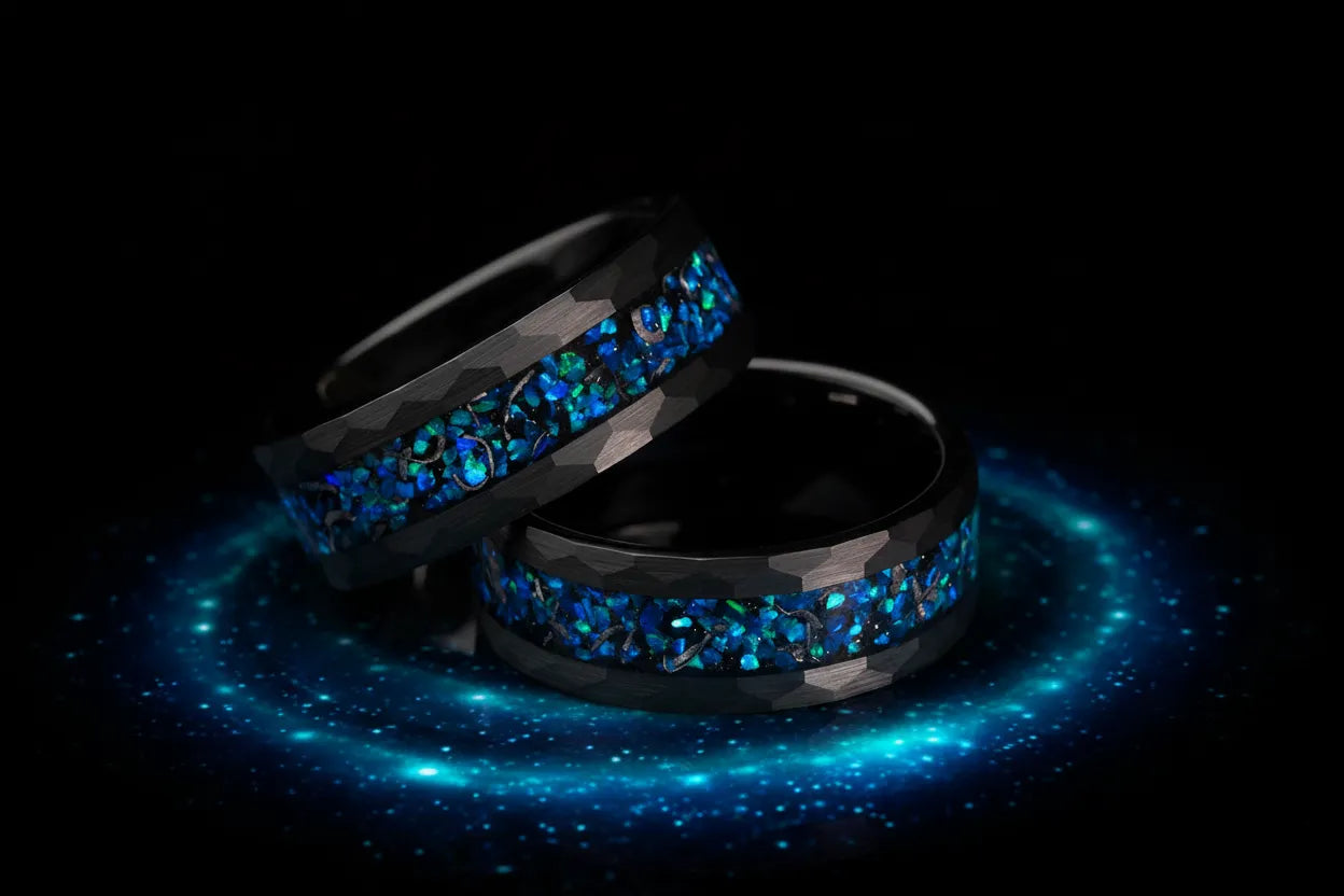 Pair of black hammered tungsten wedding bands with galaxy blue opal inlay on dark background