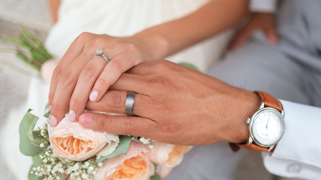 Bride and groom holding hands over a rose bouquet, showing diamond engagement ring and wedding band.