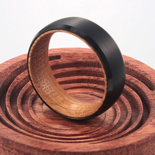 THE TIBERIUS men's wedding band: Matte black tungsten carbide ring with oak wood interior, 8mm wide, on carved wood stand.