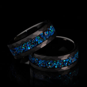 Two black tungsten rings with faceted hammered design and blue opal inlay.
