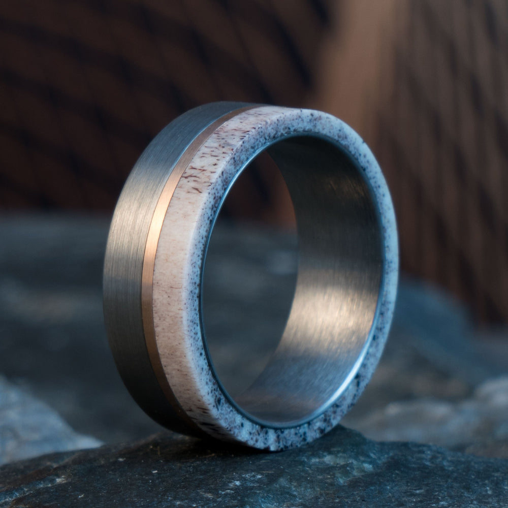 Brushed silver tungsten ring with rose gold inlay and natural deer antler band on rock.